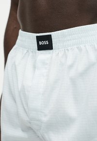 Gros plan sur le torse d'un homme à la peau foncée portant un boxer blanc à taille élastique avec une étiquette noire « BOSS » au centre de la ceinture.