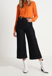 Orange langærmet bluse parret med højtaljede, vide denimjeans i mørk farve og hvide hæle støvler. Rene og glatte stofstrukturer.