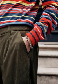 Triibuline kampsun punases, kollases, sinises ja valges; oliivirohelised püksid; hõbedane kettidega käevõru; tekstureeritud materjalid; lähivõte vöökohtadest ülespoole.