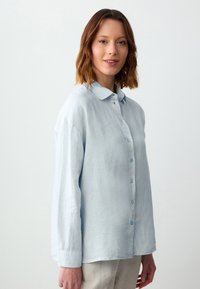 Himmelblaues Langarmhemd aus Textilgewebe, mit spitzem Kragen, durchgehender Knopfleiste und blauen Knöpfen.