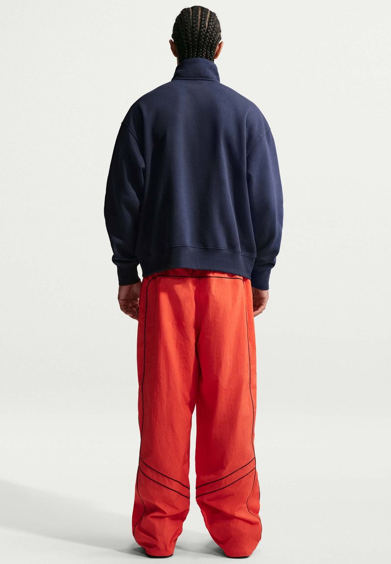 Homme aux cheveux tressés, vu de dos, portant une veste bleu marine et un pantalon ample orange vif avec des détails de coutures noirs.