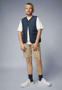 Mörkblå quiltad väst med två framfickor, bärs över en vit t-shirt, i kombination med beige shorts och vita sneakers, mot en grå bakgrund.