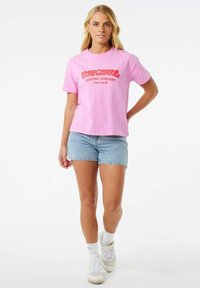 Roze katoenen T-shirt met grote rode opdruk "Ripcurl Surfing Company". Gecombineerd met denim shorts en witte sneakers, waardoor een casual look ontstaat.