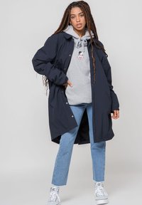 Marine lang frakk, med knapper, kombinert med en grå hettegenser med grafikk. Modellen har på seg lys blå jeans og hvite joggesko.