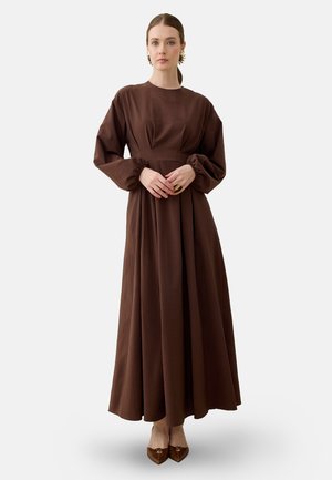 Frau mit heller Haut und braunem, zurückgebundenem Haar, die ein langes braunes Kleid mit Puffärmeln und braune spitze Schuhe trägt, steht mit gefalteten Händen.