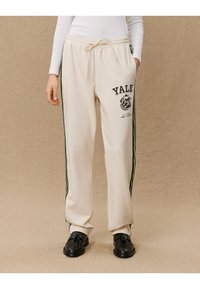 Cream-colored sweatpants with green side stripes, featuring a Yale graphic and bulldog print on the left thigh, elastic waistband, and casual fit.