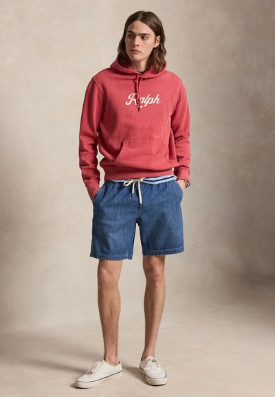 Νεαρός άνδρας με κόκκινο φούτερ με το κείμενο «Ralph», μπλε τζιν σορτς, λευκά αθλητικά παπούτσια, με τα χέρια στις τσέπες, στέκεται μπροστά σε απλό φόντο.