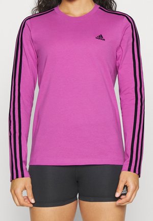 Persona con una camiseta de manga larga morada de Adidas con rayas negras en las mangas y pantalones cortos negros de Adidas.