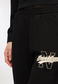 Czarne bawełniane spodnie dresowe z elastycznym paskiem w talii. Posiadają biały nadruk z logo "Brooklyn" oraz dużą literę "N" na lewym udzie.