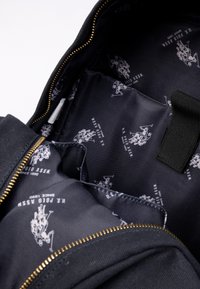 Svart ryggsäck interiör med ett dragkedjeöppning, prydd med ett slätt tygfoder dekorerat med ett vitt U.S. Polo Assn. logomönster.