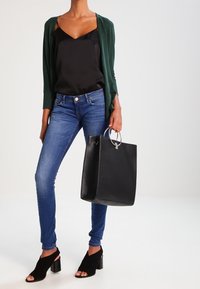 Bolso negro con un asa de anillo plateado, combinado con un cárdigan verde, una top negro y jeans ajustados azules. Zapatos de tacón abierto negros.