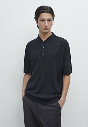 Giovane uomo con capelli scuri che indossa una polo nera a maniche corte in maglia e pantaloni neri, in piedi contro uno sfondo chiaro e uniforme.