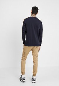 Marine blauwe sweatshirt met gouden schouderstrepen, beige cargobroek, grijze sneakers en witte sokken, gezien van achteren tegen een effen achtergrond.