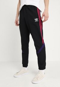 Černé sportovní kalhoty s růžovými a fialovými bočními pruhy, vyrobené z lehkého materiálu, s elastickým pasem a zúženými manžetami.