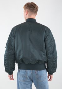 Tamnozeleni bomber jakna s sjajnim završetkom, rebrastim okovratnikom i manžetama, podstavljenim ramenima i detaljima na bočnim džepovima. Nosi se preko svijetloplavih traperica.