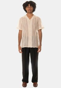 Camicia a maniche corte color crema con motivo in pizzo, dotata di colletto e bottoni, abbinata a pantaloni neri e scarpe slip-on intrecciate marroni.