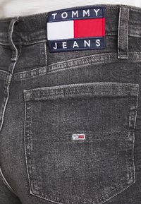 Grå denimjeans med en texturerad yta, som har en stor "TOMMY JEANS"-etikett på midjan och en liten logopatch på bakfickan.