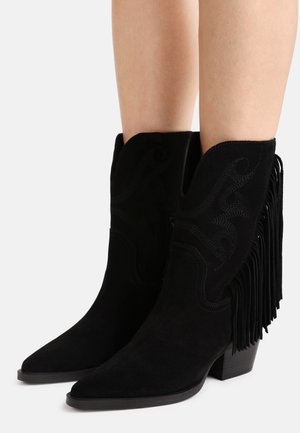 Stivali da cowboy in camoscio nero con punte appuntite, tacchi a blocco di altezza media, frange laterali e cuciture decorative indossati su gambe nude.