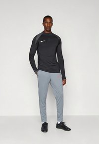 Czarny sportowy top z długim rękawem i zapięciem na suwak do ćwierć długości, szare zwężane spodnie dresowe z czerwonym logo oraz czarne buty sportowe. Gładkie, dopasowane materiały.