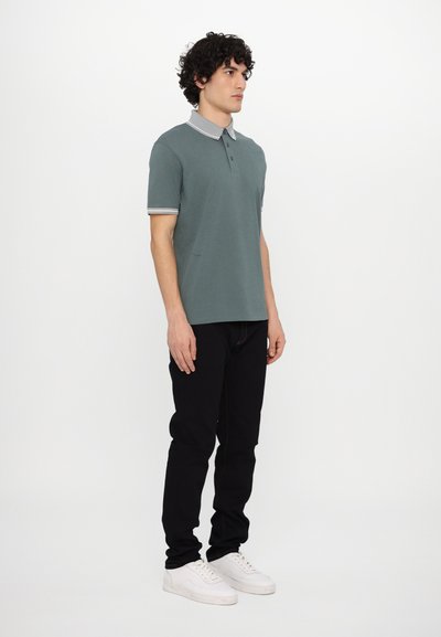 Polo skjorte i dempet teal med en kontrasterende lys grå krage og mansjetter, kombinert med svarte bukser og hvite sneakers.