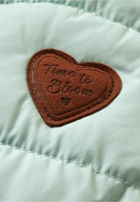 Puce en forme de cœur marron avec le texte "Time to Bloom" cousu sur un tissu matelassé vert clair.