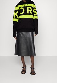 Sudadera negra con rayas amarillas y logo "KORS", combinada con una falda de cuero negra hasta la rodilla y sandalias negras con hebillas.