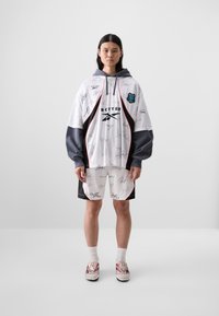 Oversized white hoodie with gray sleeves, featuring black signatures and a logo. Paired with black shorts and red-accented sneakers.