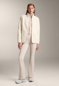 Giacca in finta pelliccia color crema con colletto alto, chiusura con zip e tasche laterali. Abbinata a un top aderente color beige chiaro e pantaloni a palazzo. Sneakers grigie.