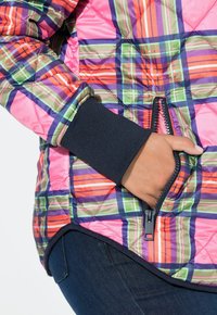 Hånd i den lynlåse lomme på en kviltet jakke med farverigt ternet mønster og sort ribkant, båret med mørkeblå jeans.