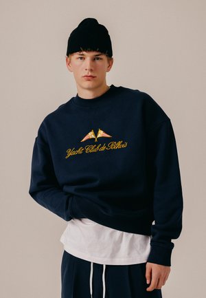 Joven con gorro negro y sudadera azul marino con texto amarillo "Yacht Club de Blkvis" y dos banderas náuticas, de pie frente a un fondo beige.