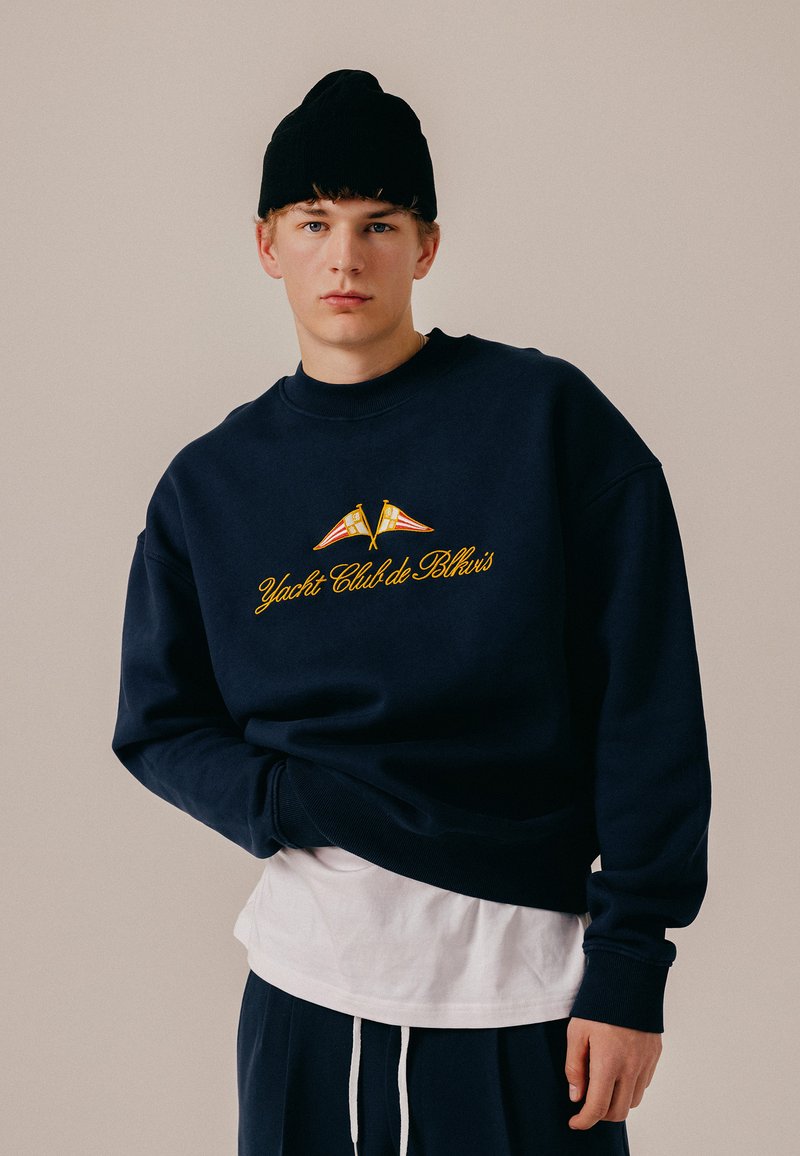 Jeune homme portant un bonnet noir et un sweat-shirt bleu marine avec le texte jaune « Yacht Club de Blkvis » et deux pavillons nautiques, debout devant un fond beige.