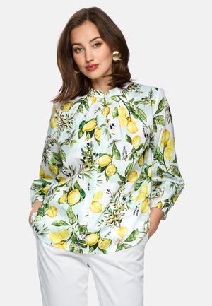 Woman wearing a long-sleeve blouse with yellow lemon and green leaf print, paired with white pants and gold round earrings.
