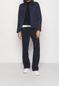 Giacca a vento blu navy con dettagli cuciti, abbinata a un maglione blu navy a trama, una t-shirt bianca e pantaloni blu navy larghi. Sneakers bianche.
