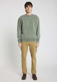 Sweatshirt vert avec un col rond, des poignets et un ourlet côtelés ; associé à un pantalon kaki et des chaussures marron ; le mannequin se tient devant un fond neutre.