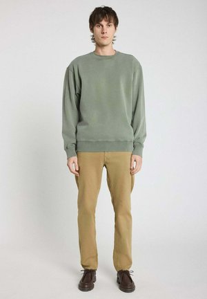 Sweatshirt vert avec un col rond, des poignets et un ourlet côtelés ; associé à un pantalon kaki et des chaussures marron ; le mannequin se tient devant un fond neutre.