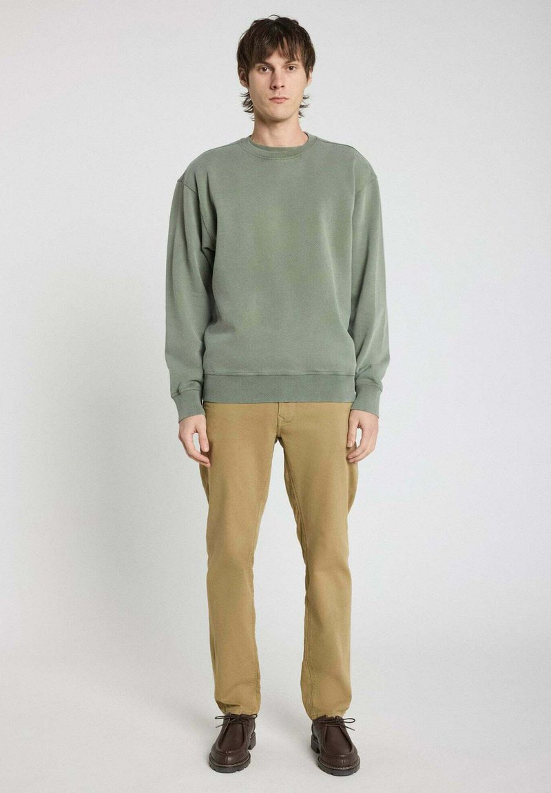 Sweatshirt vert avec un col rond, des poignets et un ourlet côtelés ; associé à un pantalon kaki et des chaussures marron ; le mannequin se tient devant un fond neutre.
