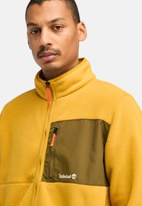 Gelbe Fleecejacke mit einer dunkelgrünen Brusttasche mit Reißverschluss und einem orangefarbenen Reißverschlussanhänger, besticktes Timberland-Logo auf der Tasche.
