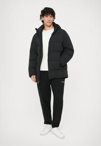 Svart puffjacka med huva, med ett quiltat mönster och dragkedjefickor, bärs över en vit skjorta och svarta träningsbyxor.