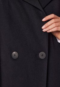 Czarny wełniany płaszcz z podwójnym zapięciem, teksturowanym materiałem, dwoma dużymi czarnymi guzikami oraz klapą w szpic. Dłoń spoczywa na materiale.