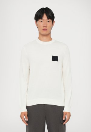 White long-sleeve sweater with a ribbed round neckline and a textured black patch on the left chest. Smooth fabric, casual fit.