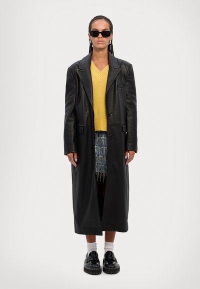 Long black leather coat over a yellow knitted sweater. Features classic collar, oversized fit, textured fabric, and chunky black platform shoes.