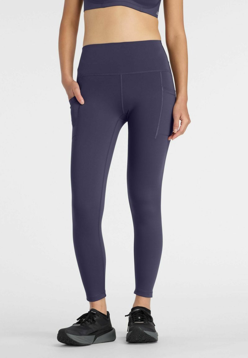 Femme portant des leggings bleu foncé taille haute avec poches latérales et chaussures de sport noires, debout devant un fond clair uni.