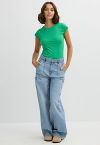 Top ajustado de manga corta verde con escote redondo, combinado con jeans cargo de pierna ancha de color azul claro y sueltos, que presentan grandes bolsillos laterales.