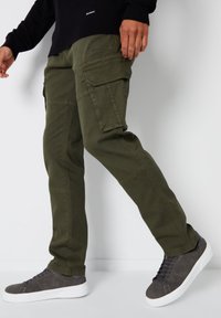 Zelené cargo kalhoty v olivové barvě s několika kapsami, rovný střih a viditelné švy. V kombinaci se šedými vysokými teniskami na neutrálním pozadí.