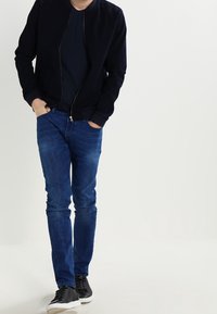 Marinblå bomberjacka över en svart tröja, matchad med blå smala jeans och svarta sneakers. Jackan har dragkedja fram och ribbade muddar.