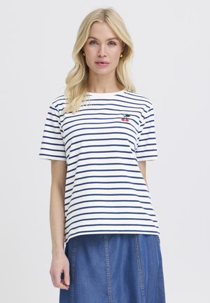 Szőke nő fehér és tengerészkék csíkos pólóban, amelyen egy kis cseresznye minta található, kék farmerruhával párosítva, fehér háttér előtt áll.