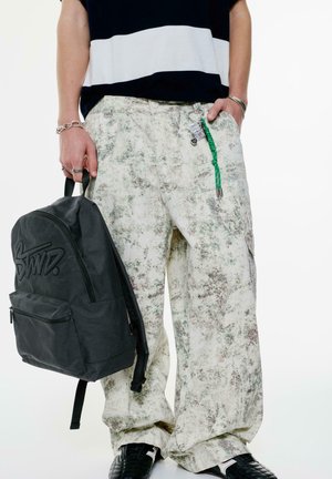 Sac à dos noir avec logo en relief, pantalons larges à motifs floraux dans des nuances de crème et de vert, et chaussures noires texturées. Détails de tenue décontractée.
