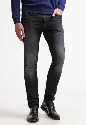 Homme portant un jean slim noir délavé avec des chaussures de ville noires brillantes, une main dans la poche, debout devant un fond uni.