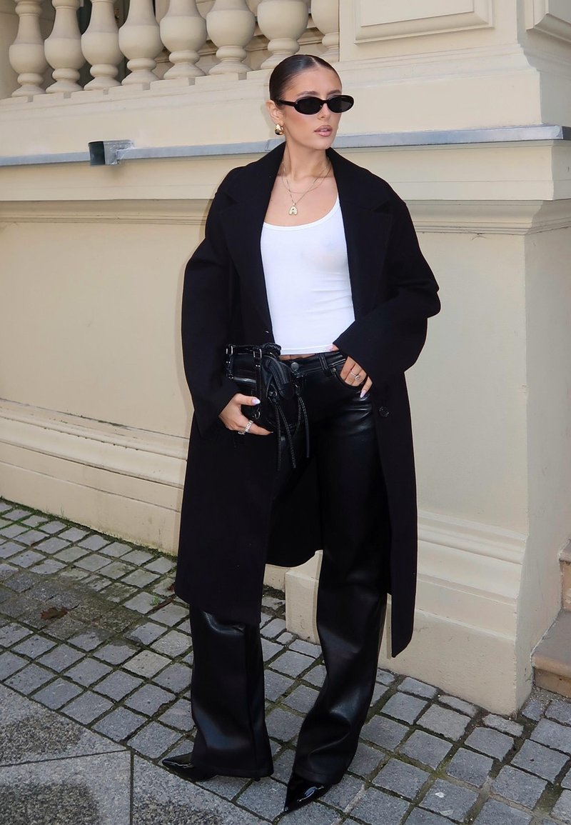 Svart oversized kappa, vit linne, svarta läderbyxor och spetsiga svarta klackar. Accessoarer inkluderar en liten svart väska och solglasögon.