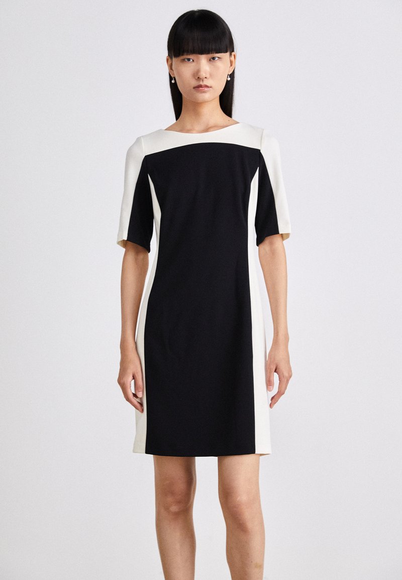 Kurzarmkleid mit schwarzem Körper und weißen Seiteneinsätzen, Rundhalsausschnitt und schmaler Silhouette. Glattes Material mit minimaler Detaillierung.